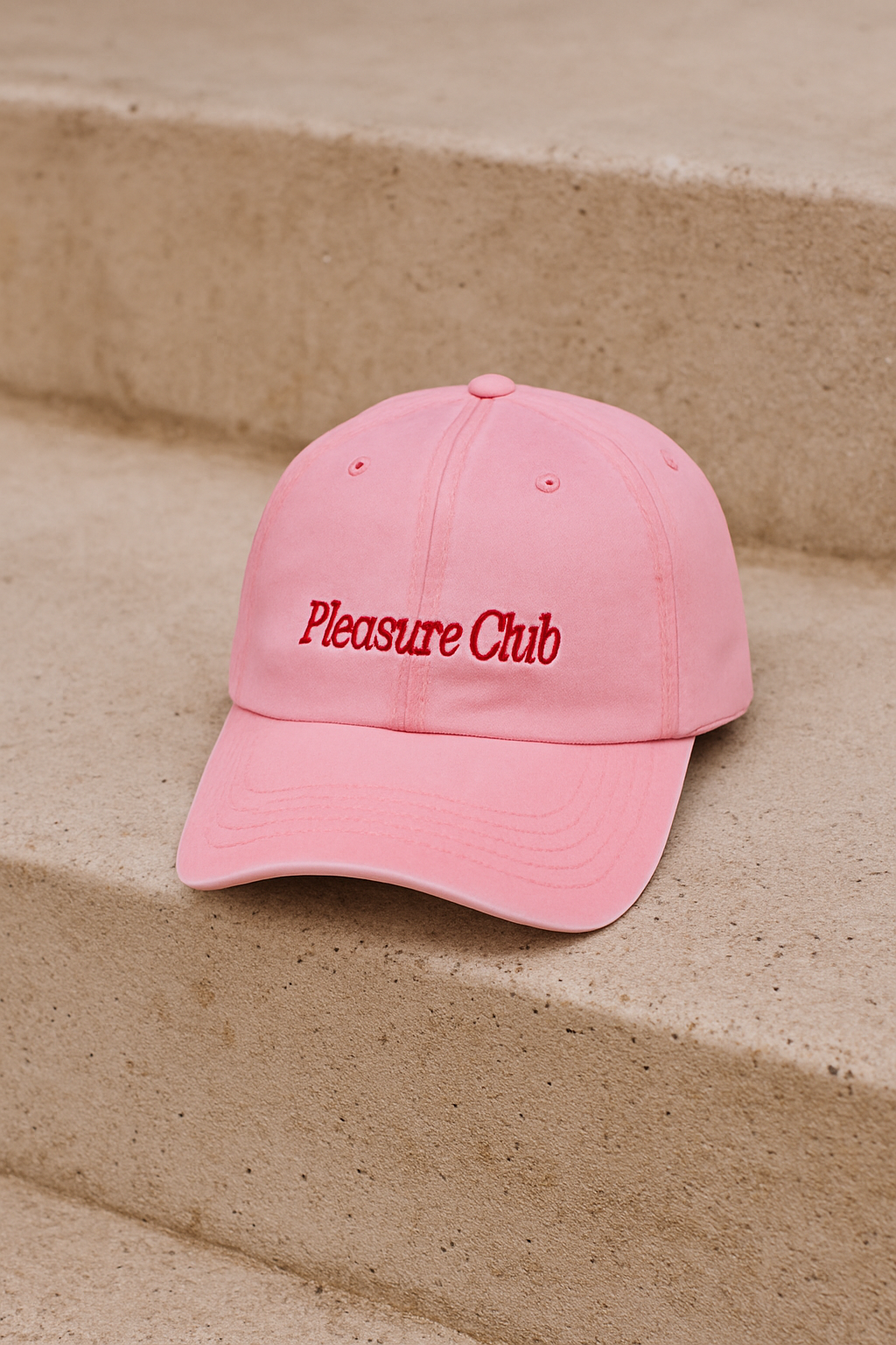 Pink cap with 'Pleasure Club' text on a beige step